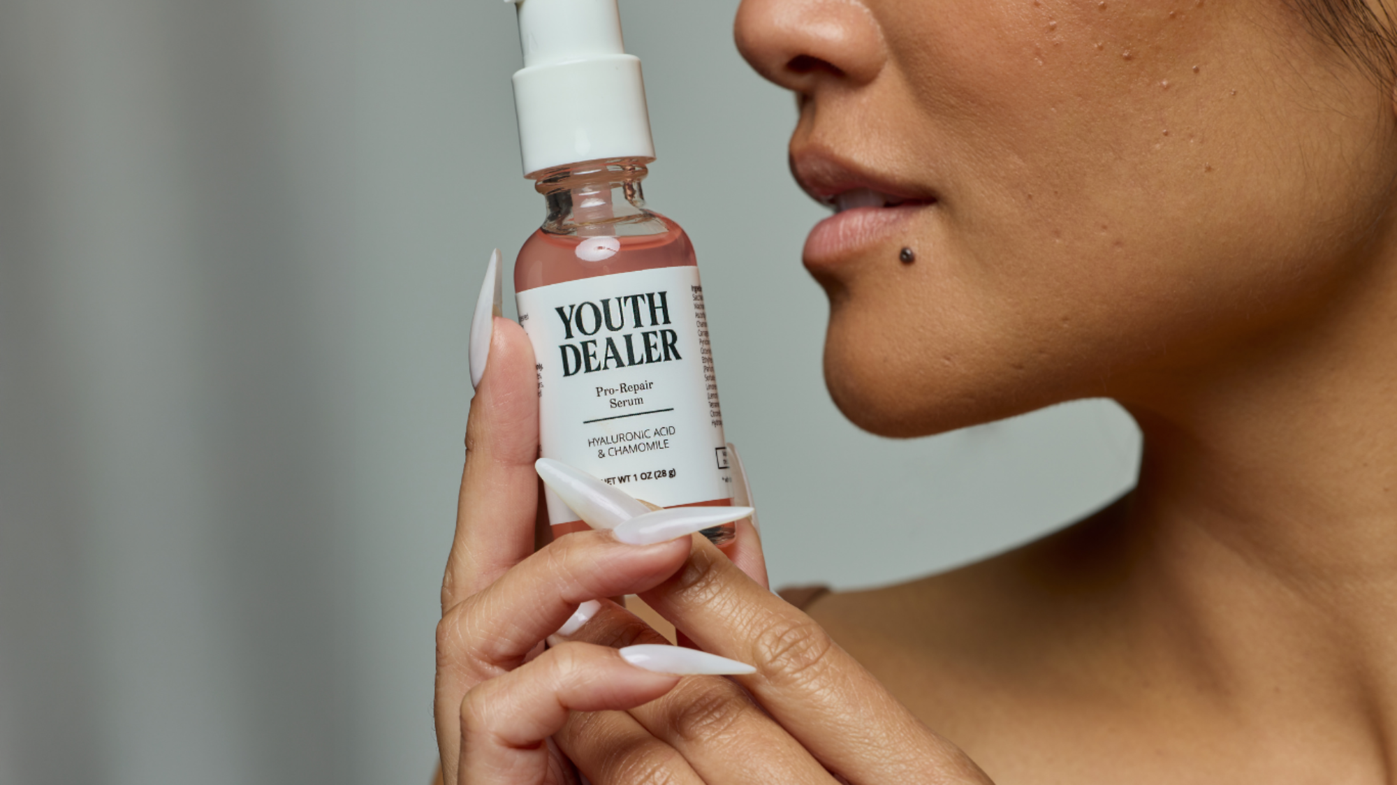Close up skin details of a woman holding a 'Youth Dealer' skincare bottle close to their face.